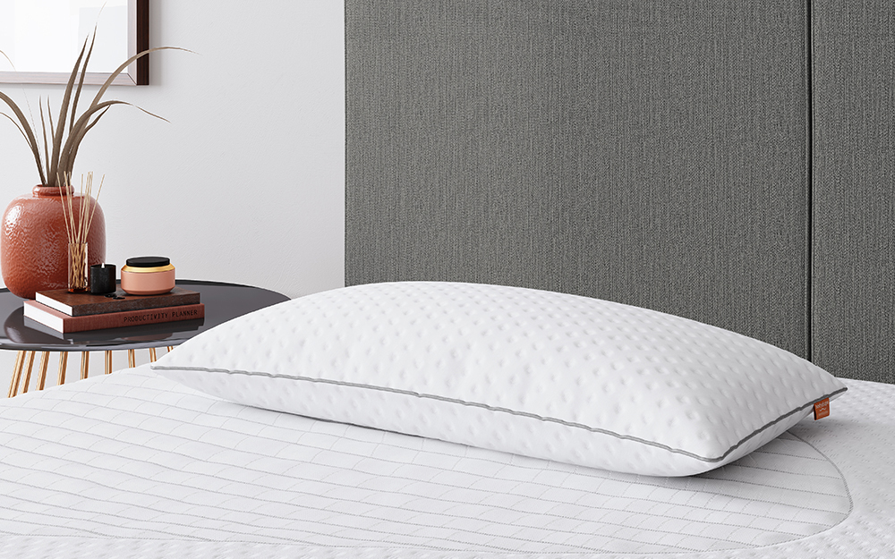 The Bodyshape Essentials Memory Foam Pillow on a grey fabric bed frame with incense sticks, books and a potted plant on the bedside table.