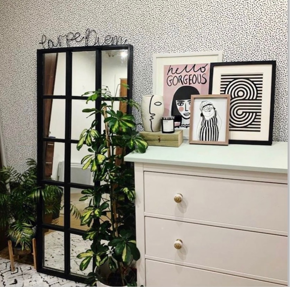 A chest of drawers next to a well decorated wall with black and white wall paper, plants and a mirror.