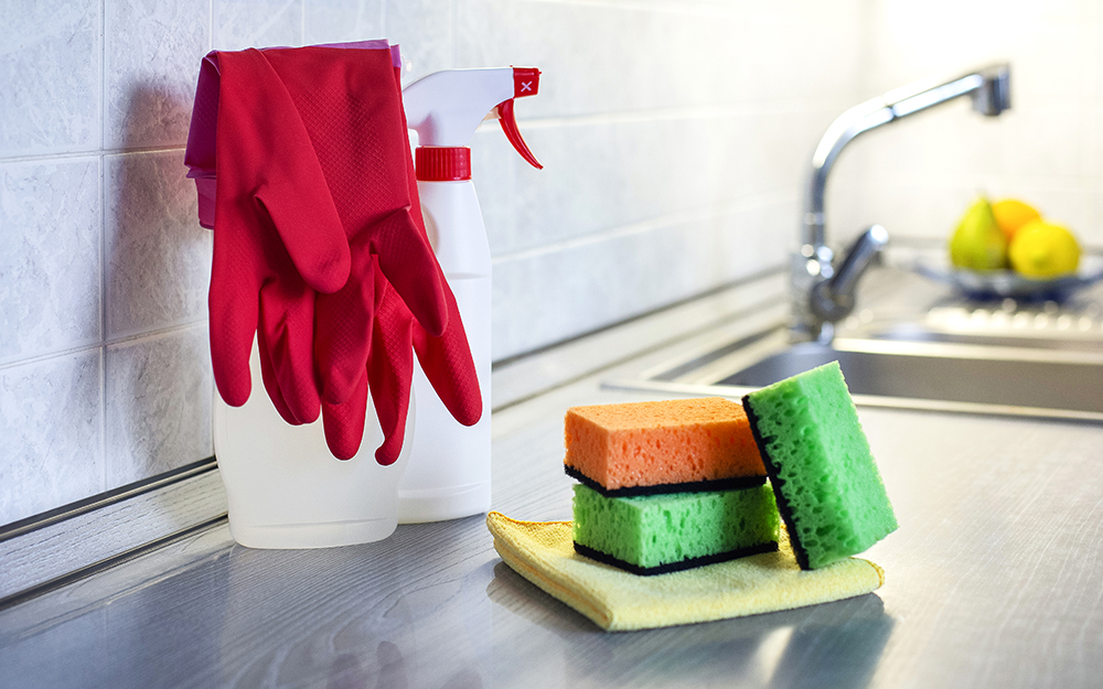 Cleaning supplies including rubber gloves, two spray bottles, sponges and clothes on a kitchen worktop next to a sink