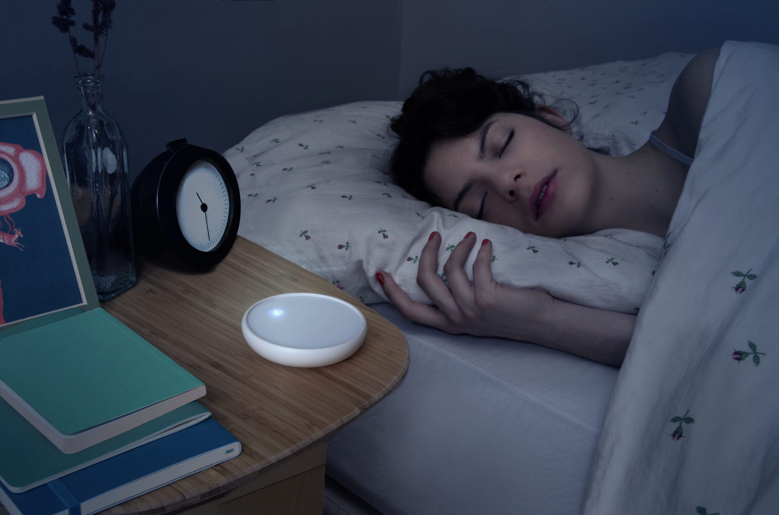 A woman sleeps soundly in bed with the Dodow beside her on a bedside table.