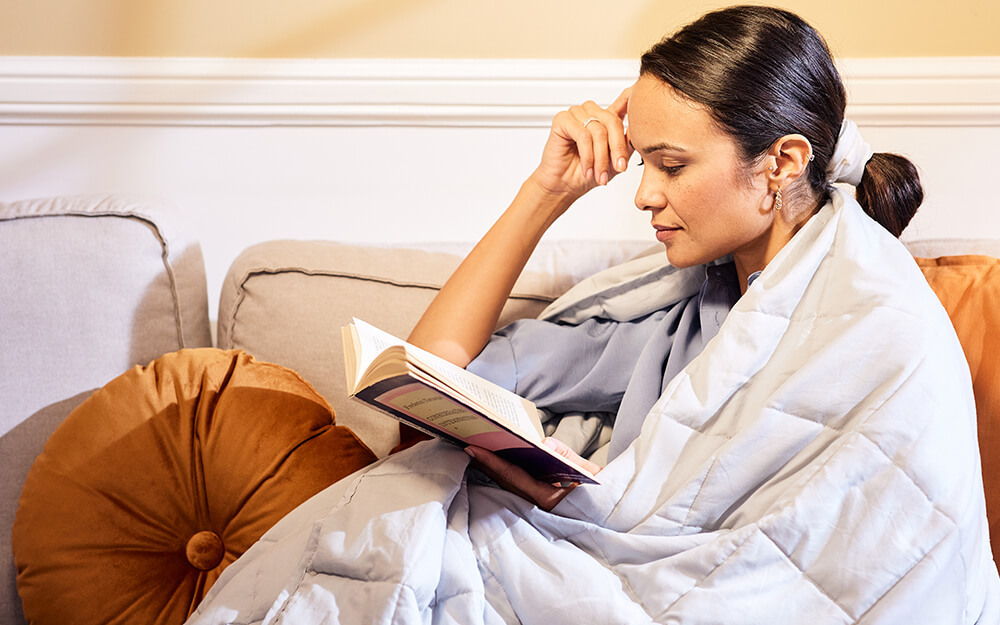 A woman sat on the sofa reading a book with the Emma Hug Weighted Blanket wrapped around her.