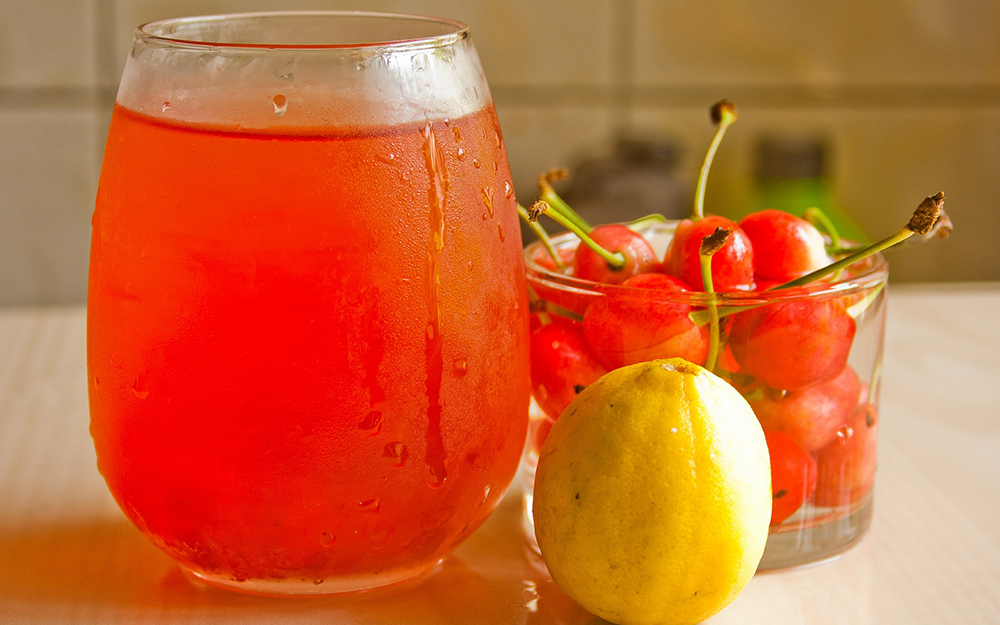 A glass of red juice next to a glass of cherries and a lemon