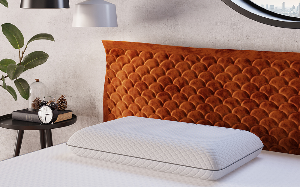 A close up of a Horizon Memory Foam Pillow on a fabric bed frame with a clock and a book on the bedside table.