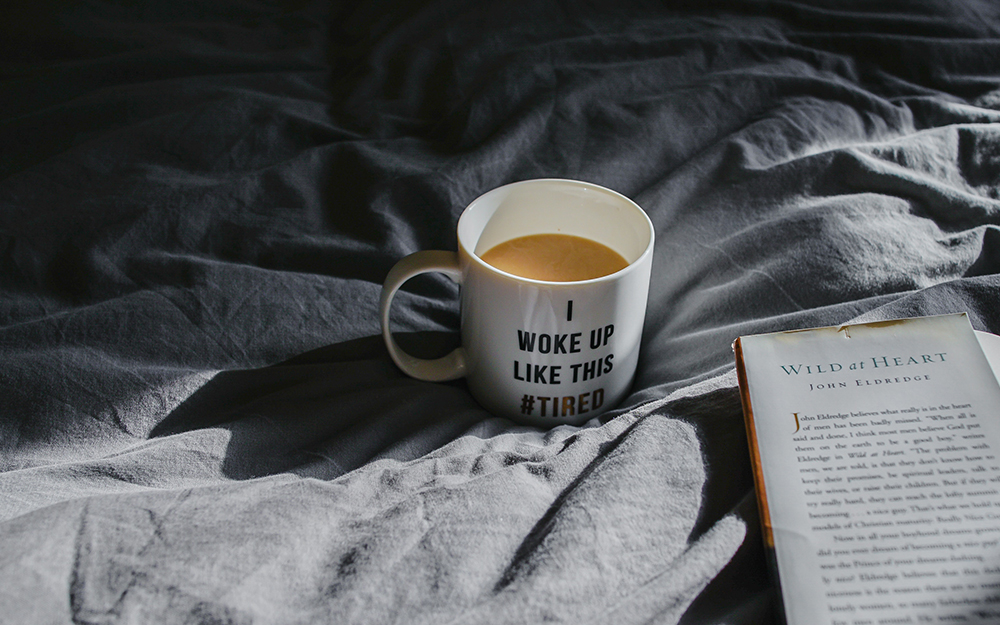 A mug that says 'I woke up like this #tired' on a bed.