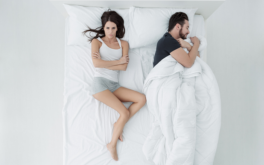 A man and a woman in bed with the man taking all of the duvet while he sleeps.