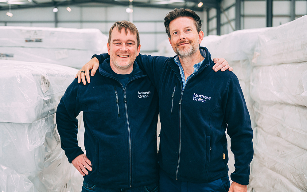 Martin Adams and Steve Adams smiling with their arms around each other in a Mattress Online warehouse.
