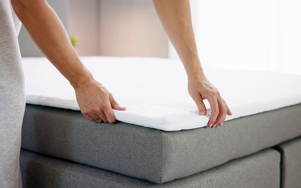 A person laying a mattress topper on top of a thin, grey mattress