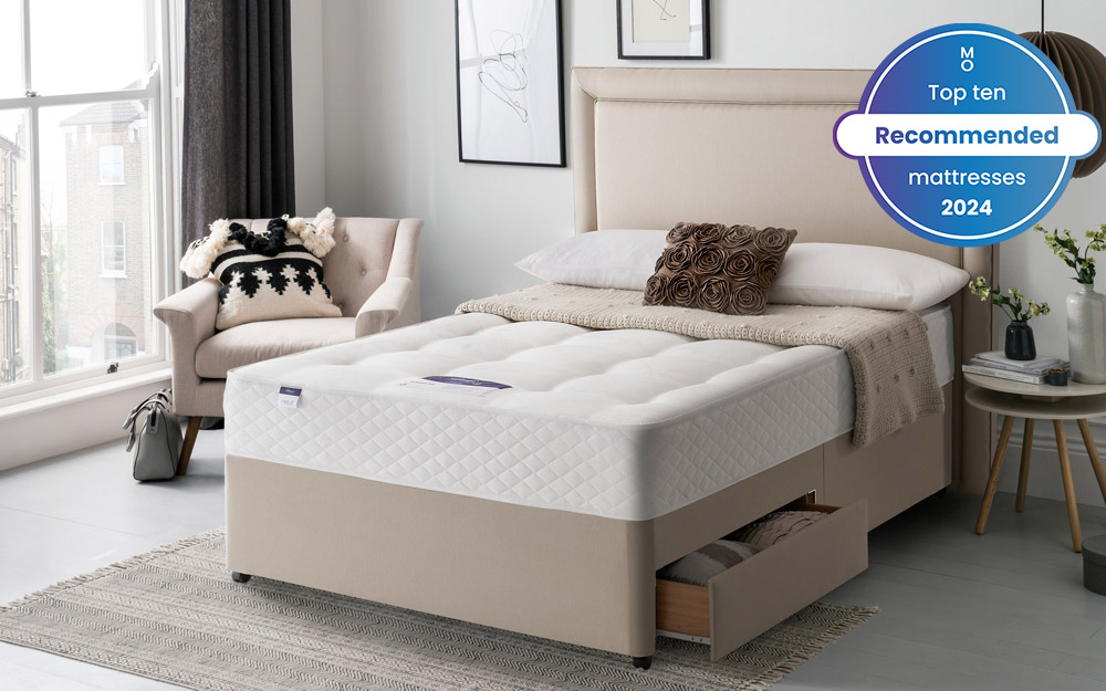 Image of the Silentnight Ortho Dream Star Miracoil Mattress on top of a beige storage bed. To the left of the bed is chair and floor length window. To the right of the bed is a bedside table. Included in the image is a badge overlay stating that this mattress is one of Mattress Online's top ten recommended mattresses of 2024.