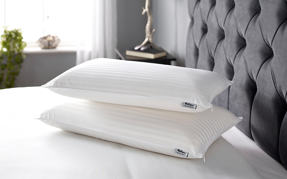 Two Relyon Superior Comfort Deep Latex Pillows stacked on top of each other on a grey fabric bed frame.