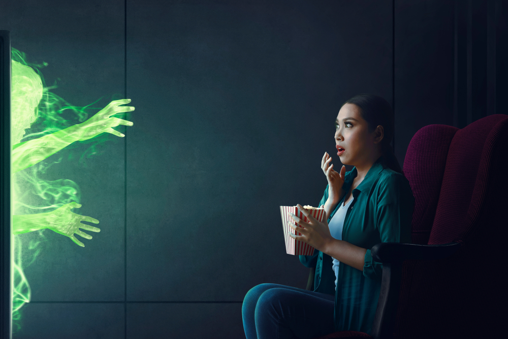 Woman looking scared watching horror film with green ghost coming from the television