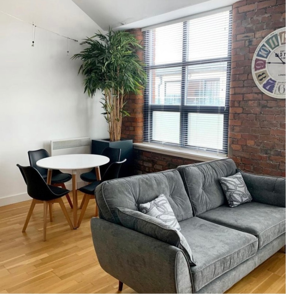 A sofa and kitchen table in an empty room next to a set of black windows and tall plant.