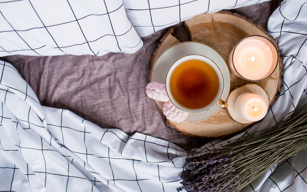 A wooden plate with a cup of tea, snacks and candles on a bed