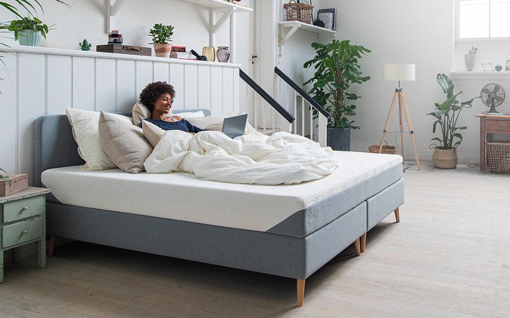 A woman sat on the TEMPUR ONE Medium Mattress in a bedroom with lots of plants in it.