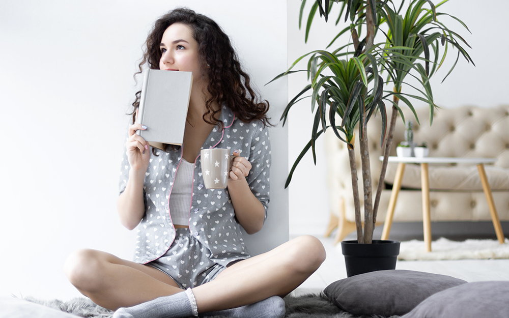 Women sitting cross legged by plant with book and mug.