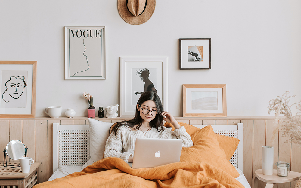 Woman reading on her laptop while sat on her double bed.